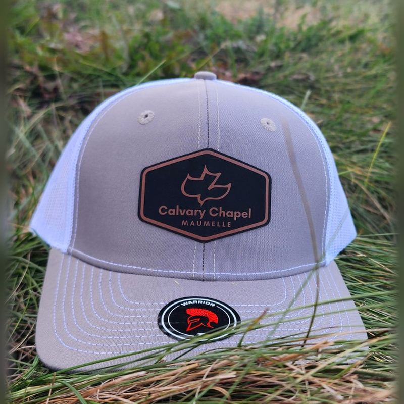 Gray and white trucker hat with a black hexagon Calvary Chapel Maumelle leatherette patch, sitting in the grass